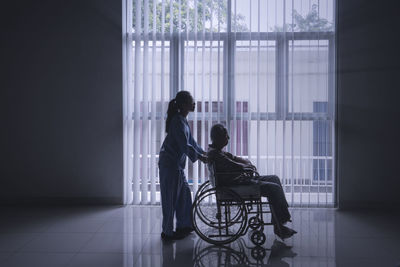 Side view of granddaughter holding wheelchair with grandfather on tied floor