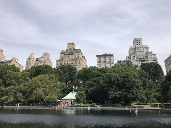 Trees by lake against buildings in city against sky