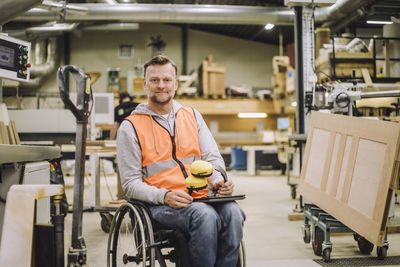 Portrait of mature carpenter with ear protectors and digital tablet sitting on wheelchair in warehouse