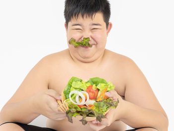 Close-up of boy holding bowl of salad making face against white background