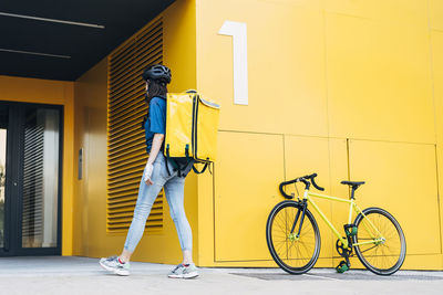 Young delivery woman with backpack walking in front of yellow wall