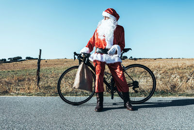 Santa claus in red clothes standing with bag near modern bicycle on empty road and looking away