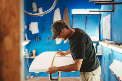 Side view of adult man measuring white board while working in small workshop with blue walls and making surf board