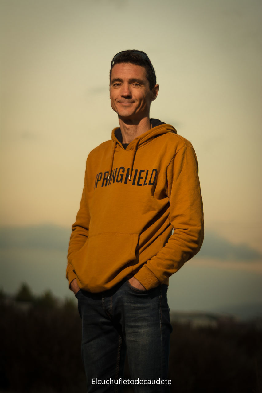 PORTRAIT OF A SMILING YOUNG MAN STANDING AGAINST SKY DURING SUNSET
