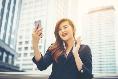 Smiling young businesswoman video conferencing against buildings in city