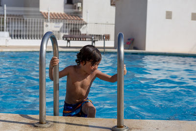 A young boy grips a ladder while getting out of a sunlit pool.