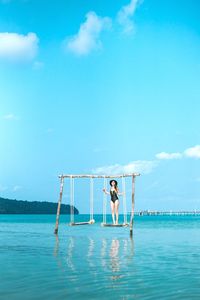 Woman standing on swing over sea against blue sky