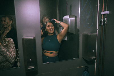 Happy young woman enjoying with friends while looking at mirror reflection in nightclub bathroom