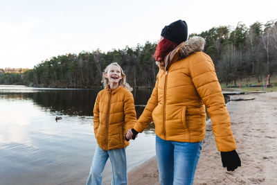 Content mother and teen girl holding hands and walking along pond in woods while enjoying weekend in autumn