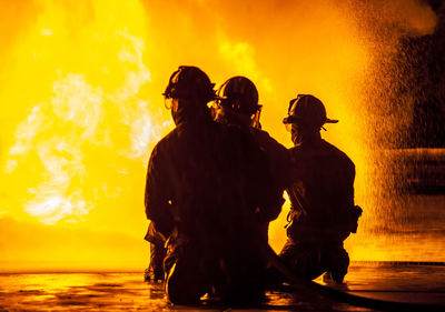 Rear view of silhouette firefighters crouching against fire at night