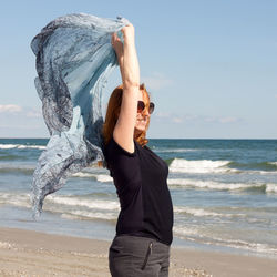 Woman with arms raised holding fabric while standing at beach against sky