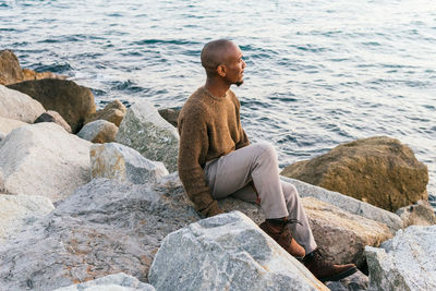 Side view full body of calm african american male looking into distance with pensive face while sitting on rocky coast near sea