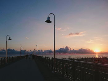Street lights on pier by sea against sky during sunset