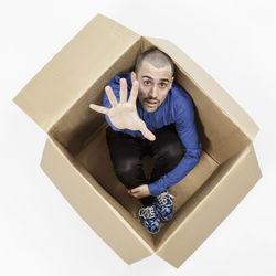 High angle portrait of man gesturing while sitting in cardboard box on white background