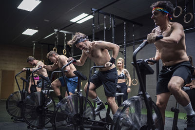 Men exercising on fitness bike with women standing in background at gym