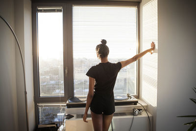 Rear view of woman looking through window while standing at home