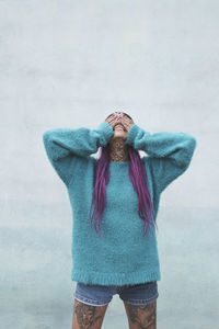 Trendy female with pink hair and tattoos on legs standing near shabby building in urban area and covering face