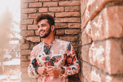 Smiling young man looking away while standing against brick wall