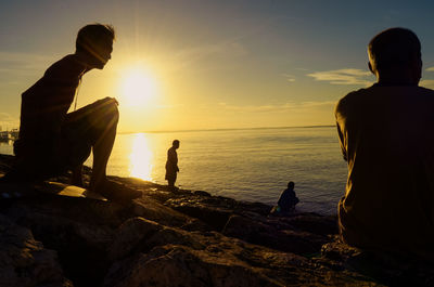 People sitting on rock by sea against sky during sunset