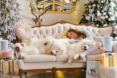 Little cute girl sitting on the sofa with two samoyed dogs