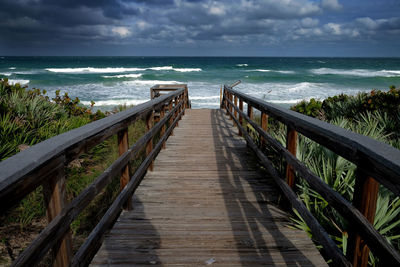 Wooden footbridge leading towards sea against sky