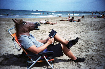 Man in horse head reading book on beach