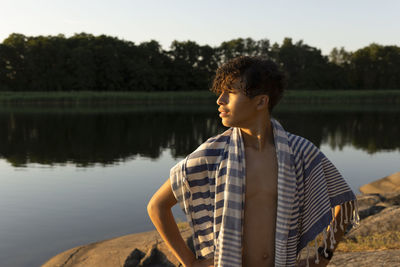 Teenage boy with towel looking away against lake during sunset
