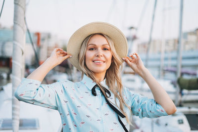 Beautiful smiling blonde young woman in blue dress and straw hat at pier in sunset time