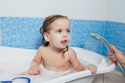 Cropped hand of mother showering shirtless daughter sitting in bathtub