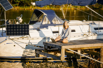 Thoughtful woman with laptop sitting on pier by moored yacht at sunny lakeside
