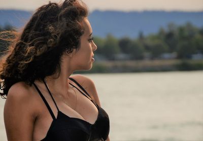 Young woman wearing bikini looking away while sitting by sea during sunset