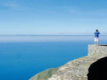 Rear view of man standing on retaining wall looking at sea against sky