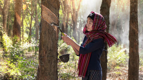 Side view of woman collecting latex while standing by tree trunk in forest