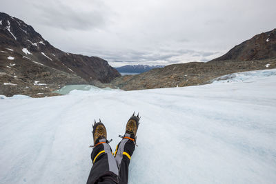 Low section of person wearing snow boots on snow covered mountains against cloudy sky