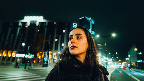 Portrait of young woman standing against illuminated city at night