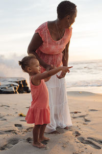 Mother with daughter standing at beach against sky during sunset