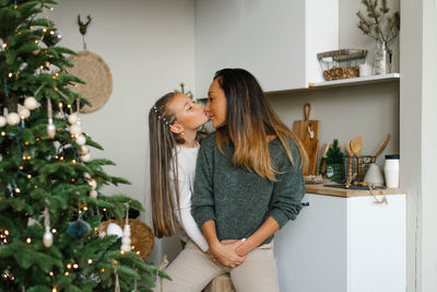 Daughter kisses mom in anticipation of christmas and new year near the christmas tree