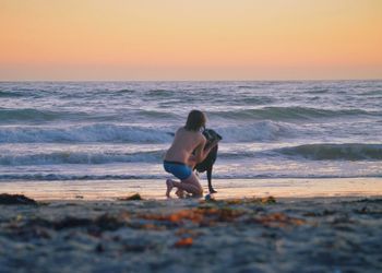 Rear view of shirtless teenage boy with dog at beach during sunset