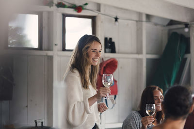 Happy young woman standing with white wine by friends at home