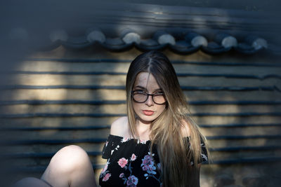 Portrait of beautiful young woman sitting by wall outdoors
