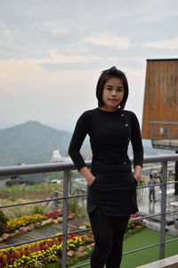 Portrait of smiling young woman with hands in pockets standing against sky