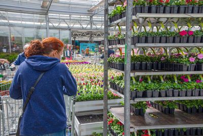 A young woman in mask chooses and buys seedlings in a garden center whith plants