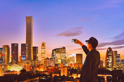 Rear view of man photographing cityscape against sky during sunset