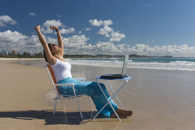 Side view of mid adult woman with arms raised with laptop sitting at beach against sky