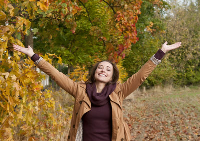 Woman with arms raised standing against trees in park during autumn