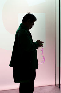 Side view of silhouette man using mobile phone while standing by wall