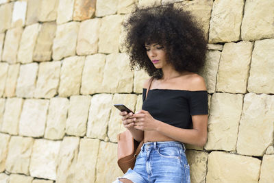 Woman with curly hair using mobile phone against wall