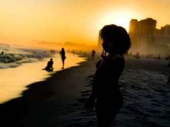 Side view of shirtless boy standing at beach against sky during sunset