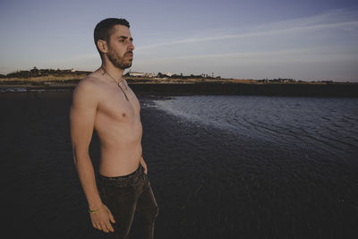 Young shirtless man looking at sea against sky