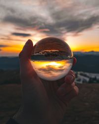 Close-up of hand holding crystal ball against sky during sunset